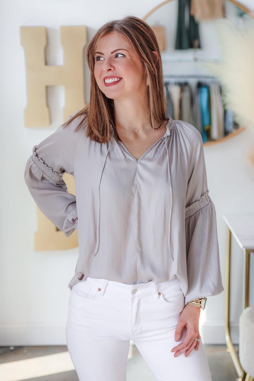 A brunette woman standing inside a shop wearing a gray satin top and white jeans. Her right hand is on her hip.