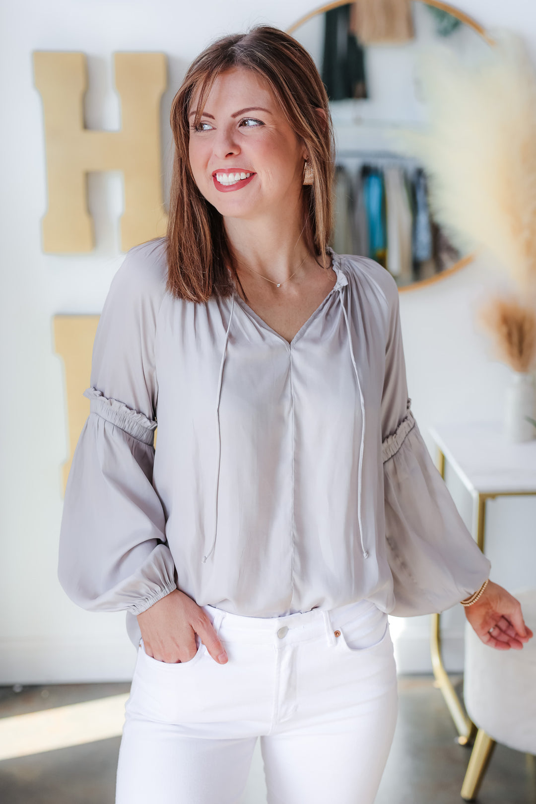 A brunette woman standing inside a shop wearing a gray satin top and white jeans. Her right hand is in her pocket.