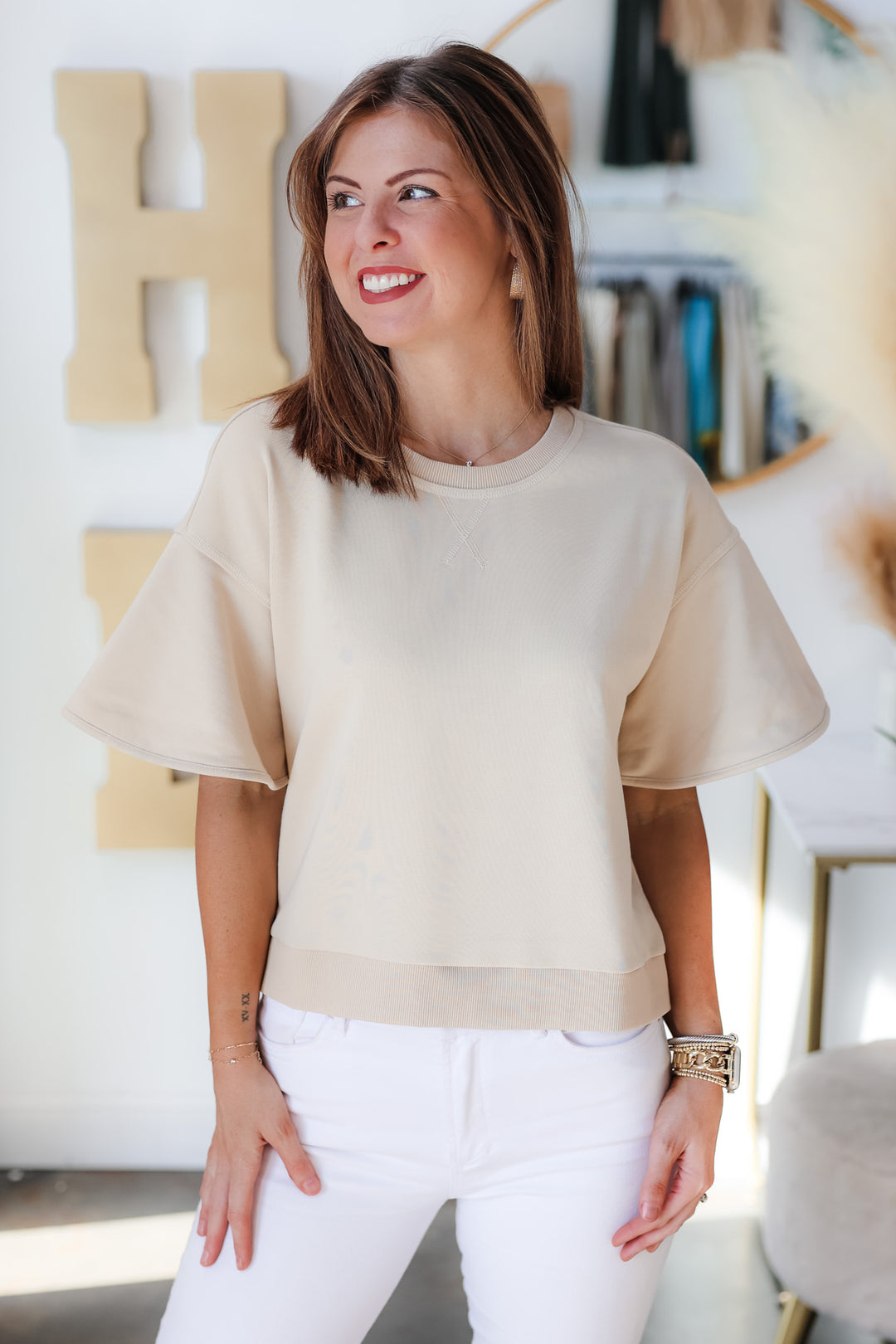 A brunette woman standing in a shop wearing a tan colored short sleeve shirt with wide sleeve and white jeans.