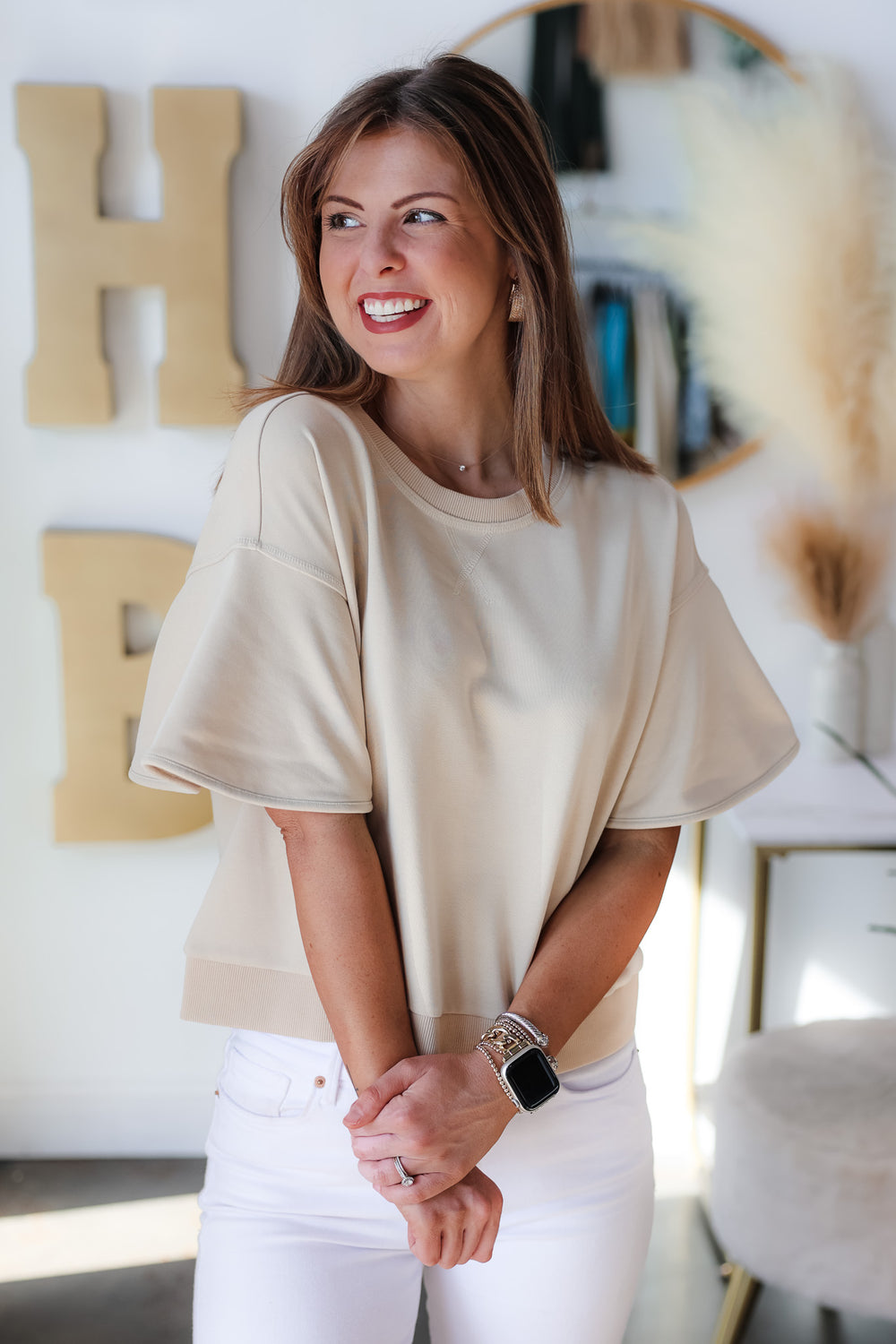 A brunette woman standing in a shop wearing a tan colored short sleeve shirt with wide sleeve and white jeans.