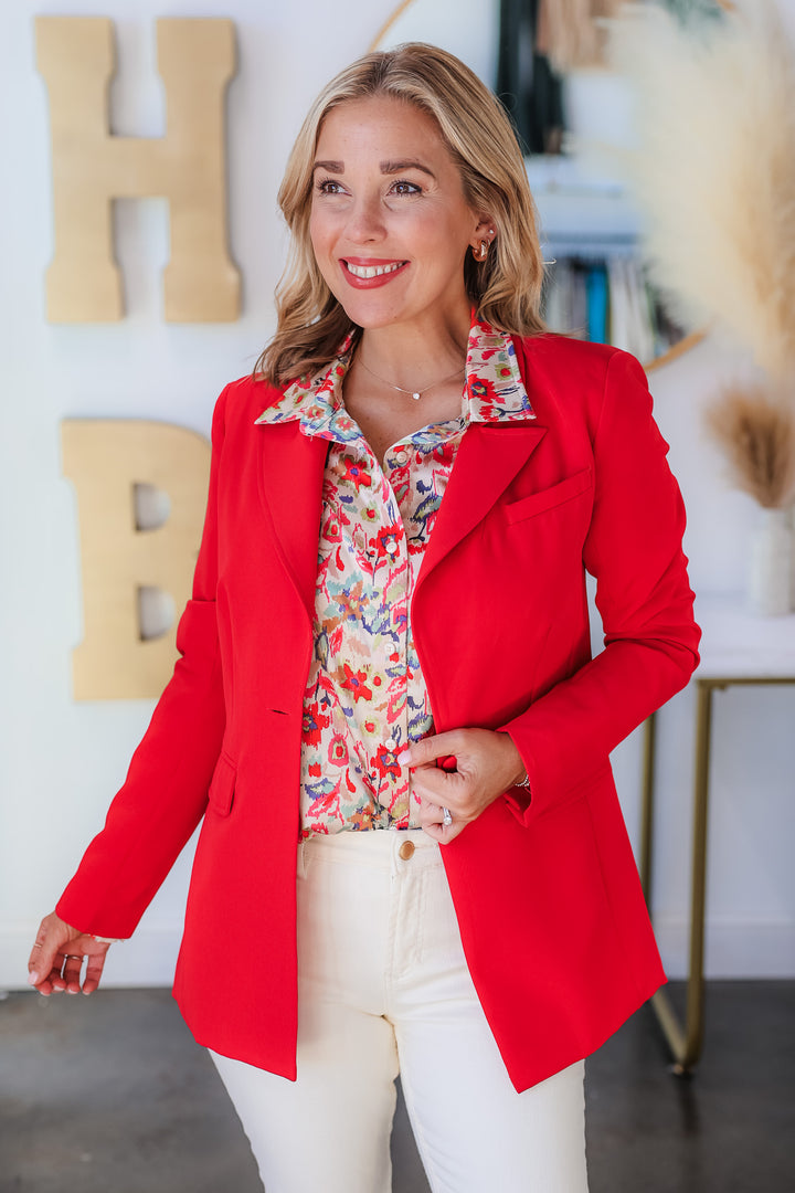 A blonde woman standing in a shop wearing a printed blouse, red blazer and ivory colored blue jeans.