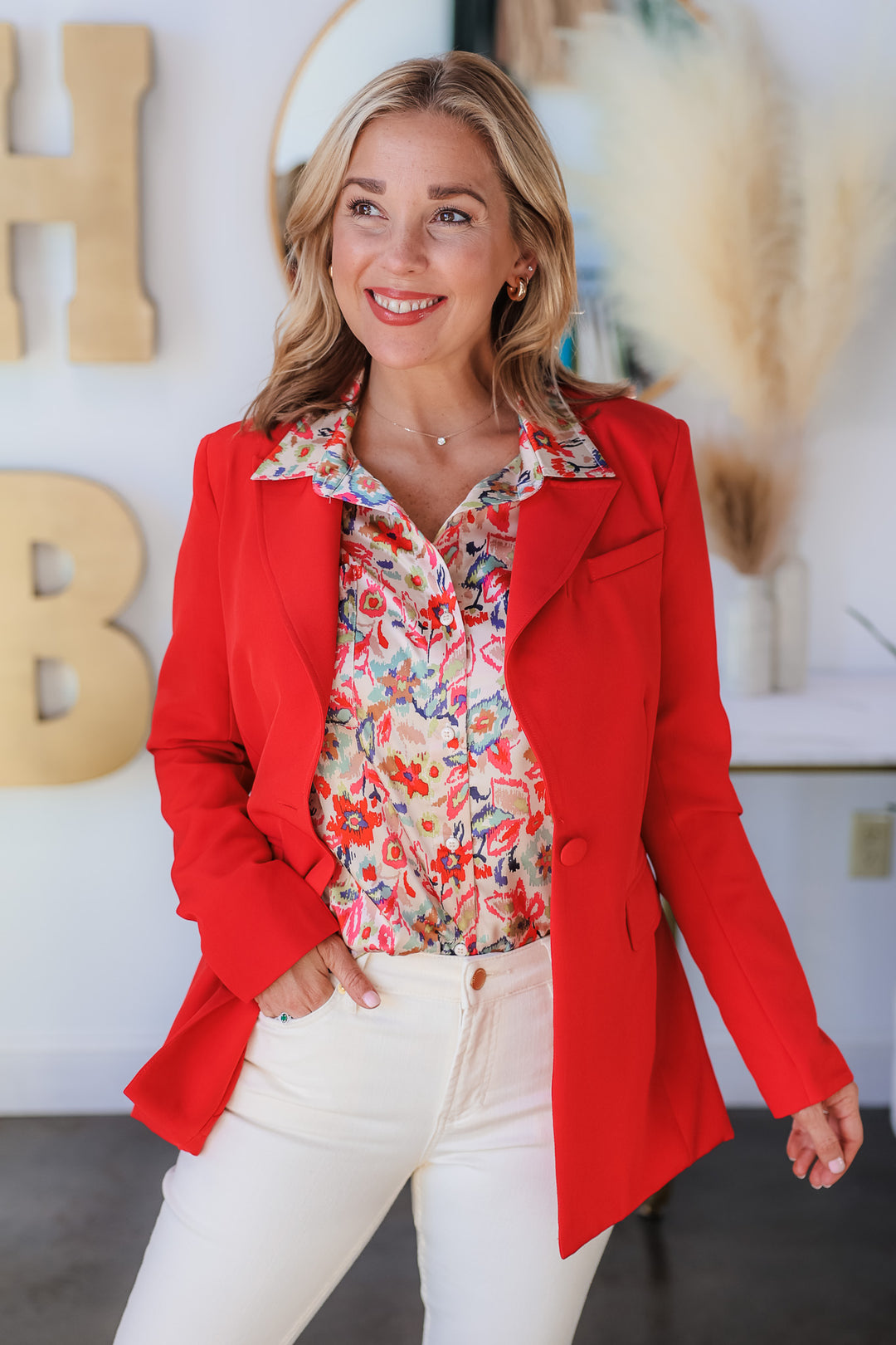 A blonde woman standing in a shop wearing a printed blouse, red blazer and ivory colored blue jeans.