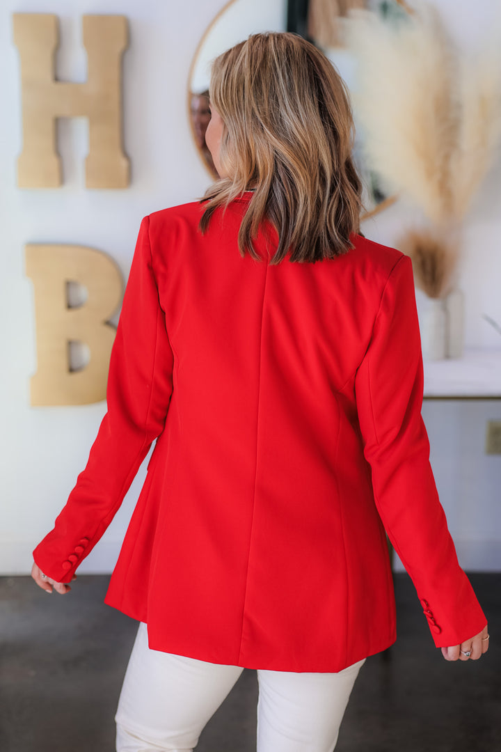 A blonde woman standing in a shop wearing a printed blouse, red blazer and ivory colored blue jeans. She is rear facing.