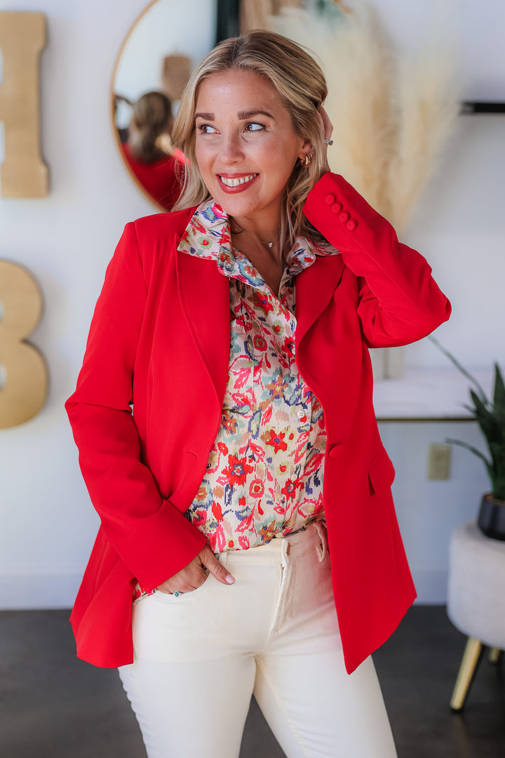 A blonde woman standing in a shop wearing a printed blouse, red blazer and ivory colored blue jeans.