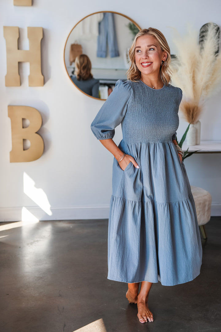 A blonde woman standing in a shop wearing a denim colored dress with 3/4 length sleeves, smocked bodice and tiered skirt.