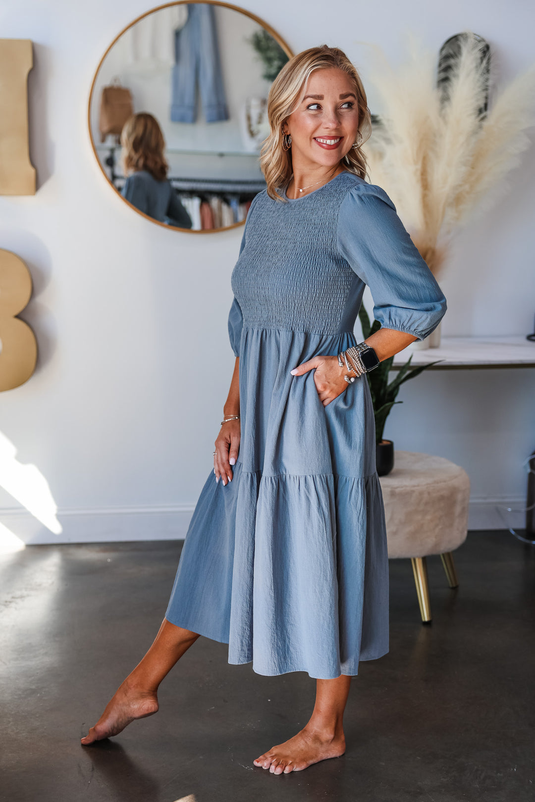 A blonde woman standing in a shop wearing a denim colored dress with 3/4 length sleeves, smocked bodice and tiered skirt.