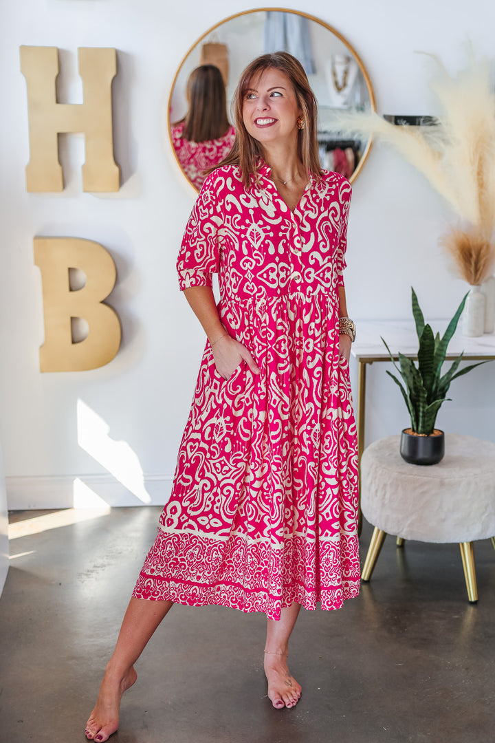 A brunette woman standing in a shop wearing a fuchsia and cream colored long dress with a button closure on top and 3/4 length sleeves.