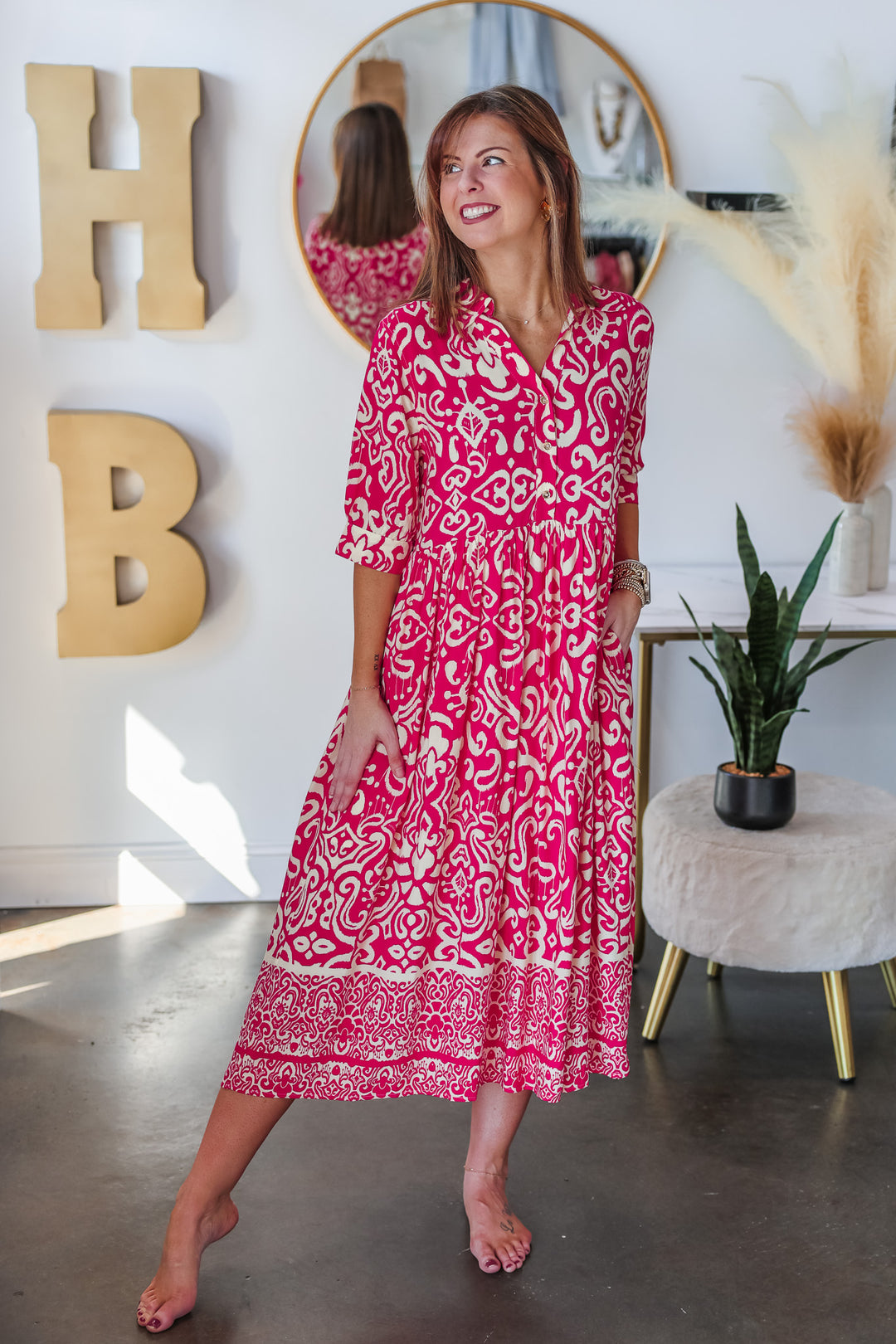 A brunette woman standing in a shop wearing a fuchsia and cream colored long dress with a button closure on top and 3/4 length sleeves.