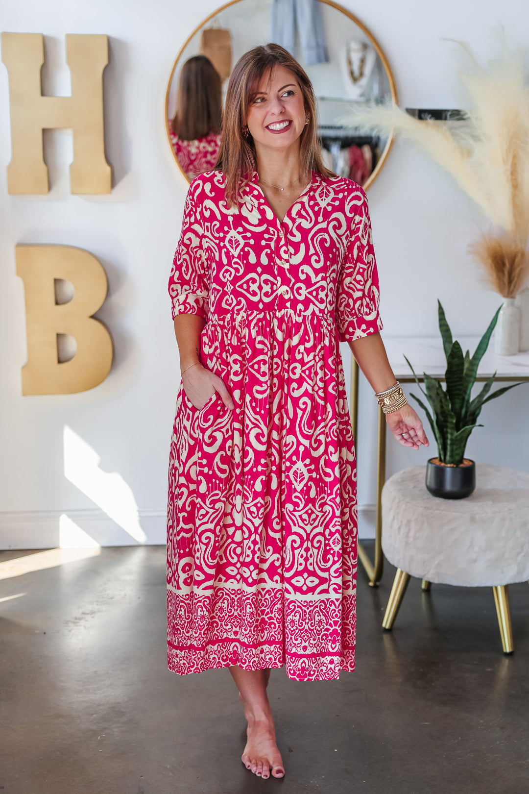 A brunette woman standing in a shop wearing a fuchsia and cream colored long dress with a button closure on top and 3/4 length sleeves.