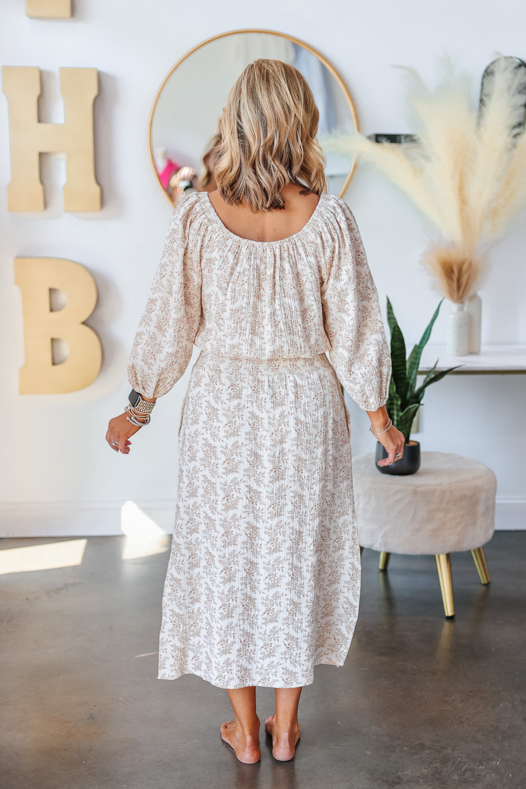 A blonde woman standing in a shop wearing a natural colored floral midi skirt with an elastic waist, side hem slits and a matching top. She is rear facing.