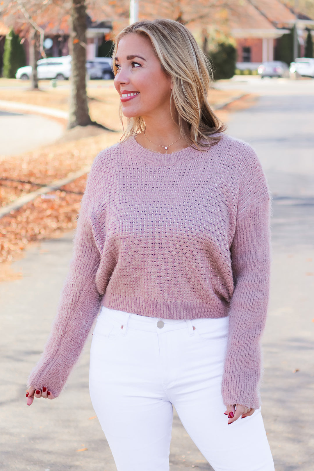 A blonde woman standing outside wearing a mauve colored long sleeve sweater and white jeans.