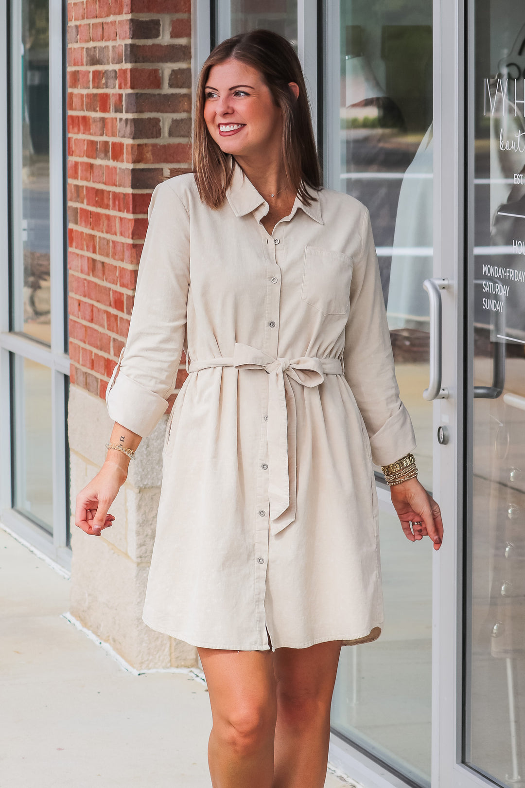 A brunette woman wearing a tan corduroy shirt dress with a tie belt. She is standing in front of a shop.