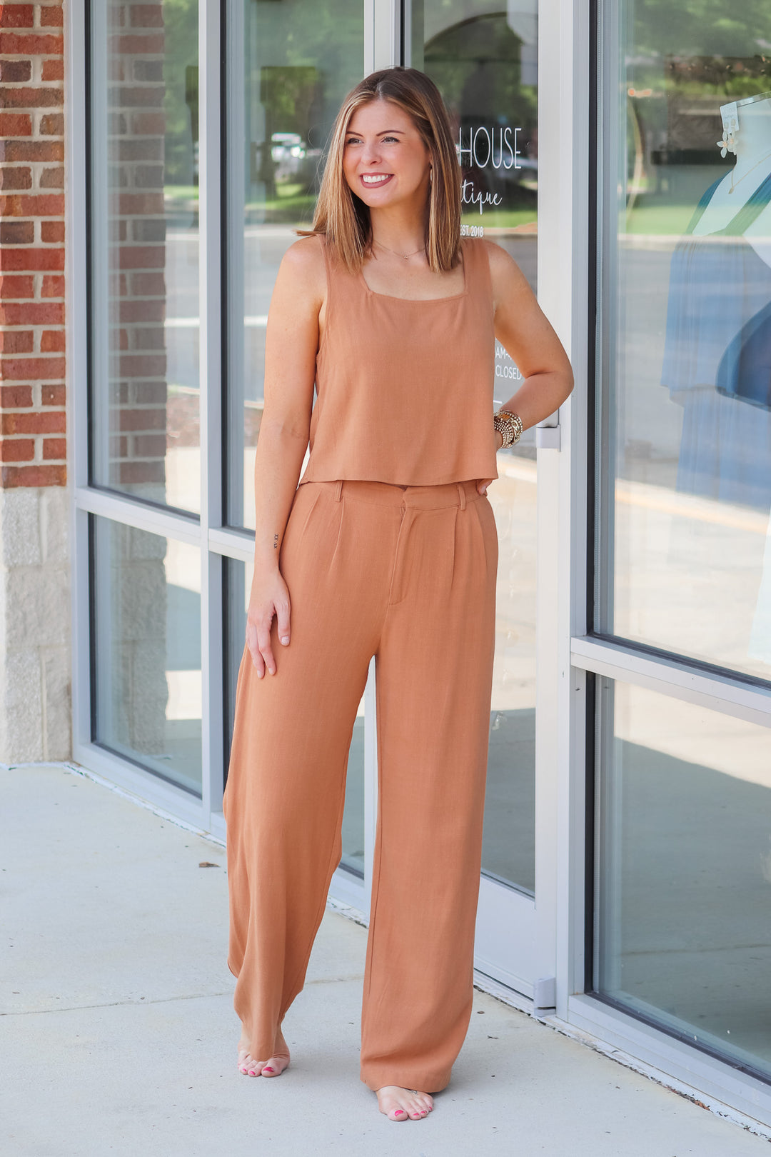 A brunette woman wearing a tan, square neckline, waist length top and matching pants.  She is standing in front of a shop. 