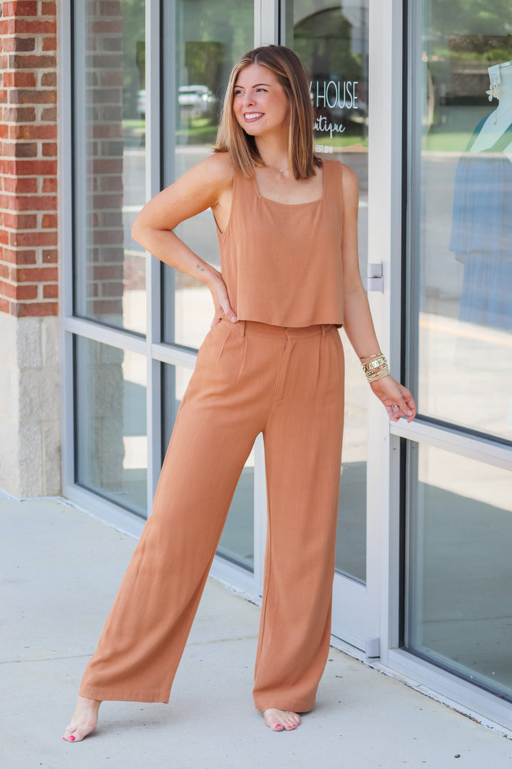 A woman wearing a tan top and tan linen pants with belt loops and pockets. She is standing in front of s shop.