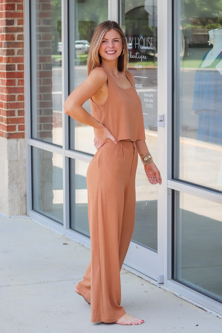 A brunette woman wearing a tan, square neckline, waist length top and matching pants. She is standing in front of a shop.  