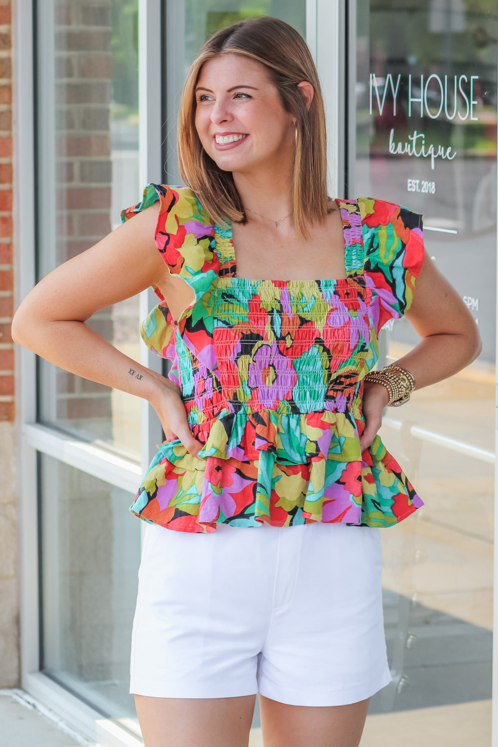 A brunette woman wearing a colorful smocked top with a ruffle peplum hem and ruffle shoulders. The colors are red, black, teal, yellow, purple and green. She is standing in front of a shop.