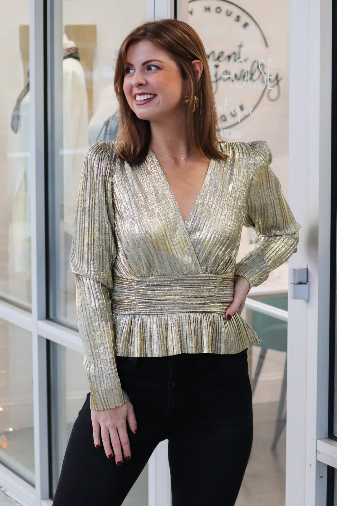 A brunette woman standing in front of a shop wearing a metallic gold pleated top with long sleeves, peplum hem and a v neck with black jeans.