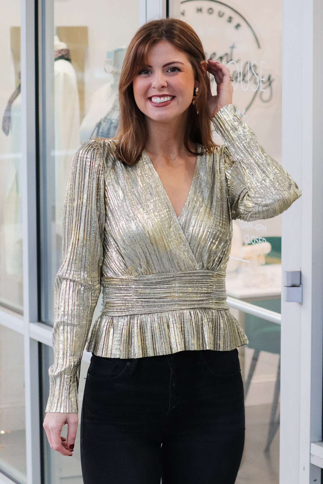 A brunette woman standing in front of a shop wearing a metallic gold pleated top with long sleeves, peplum hem and a v neck with black jeans.