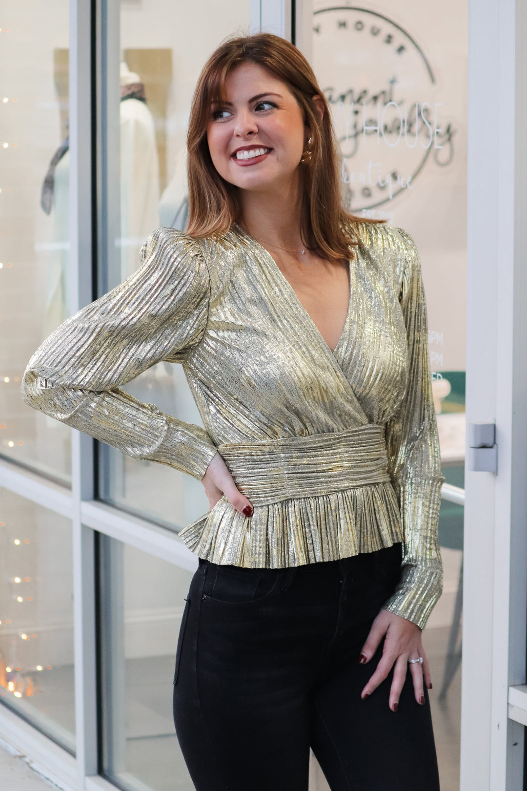 A brunette woman standing in front of a shop wearing a metallic gold pleated top with long sleeves, peplum hem and a v neck with black jeans.