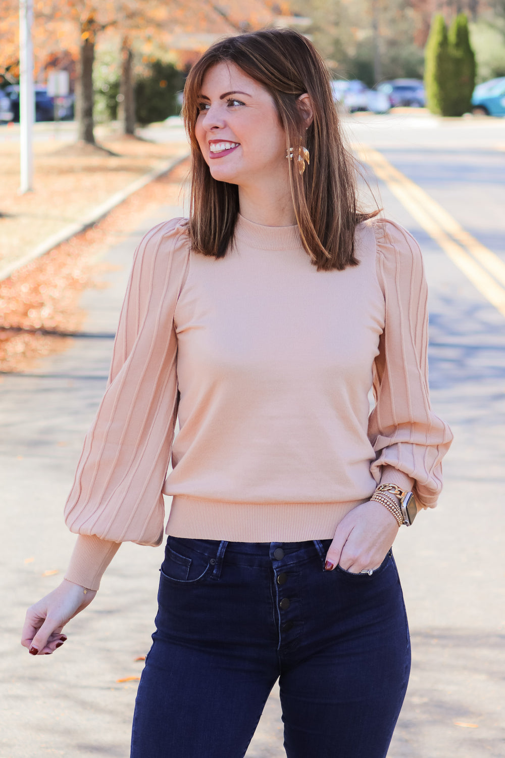 A brunette woman standing outside wearing an oat colored detailed long sleeve sweater with blue jeans.