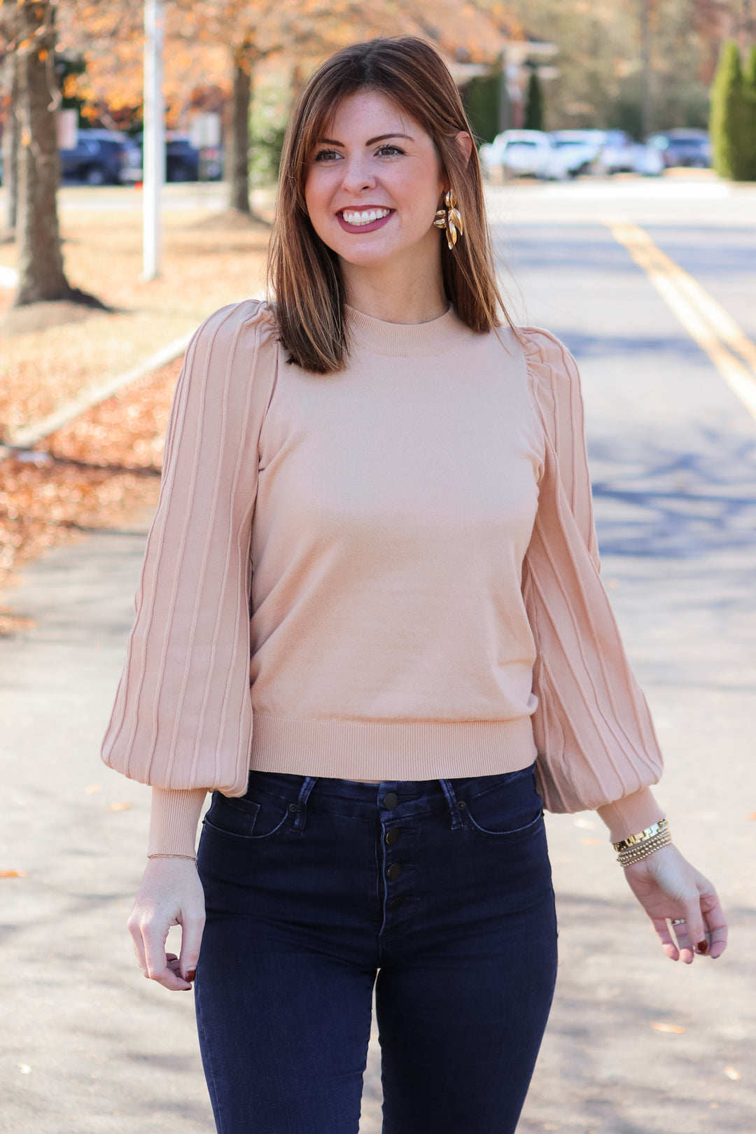 A brunette woman standing outside wearing an oat colored detailed long sleeve sweater with blue jeans.