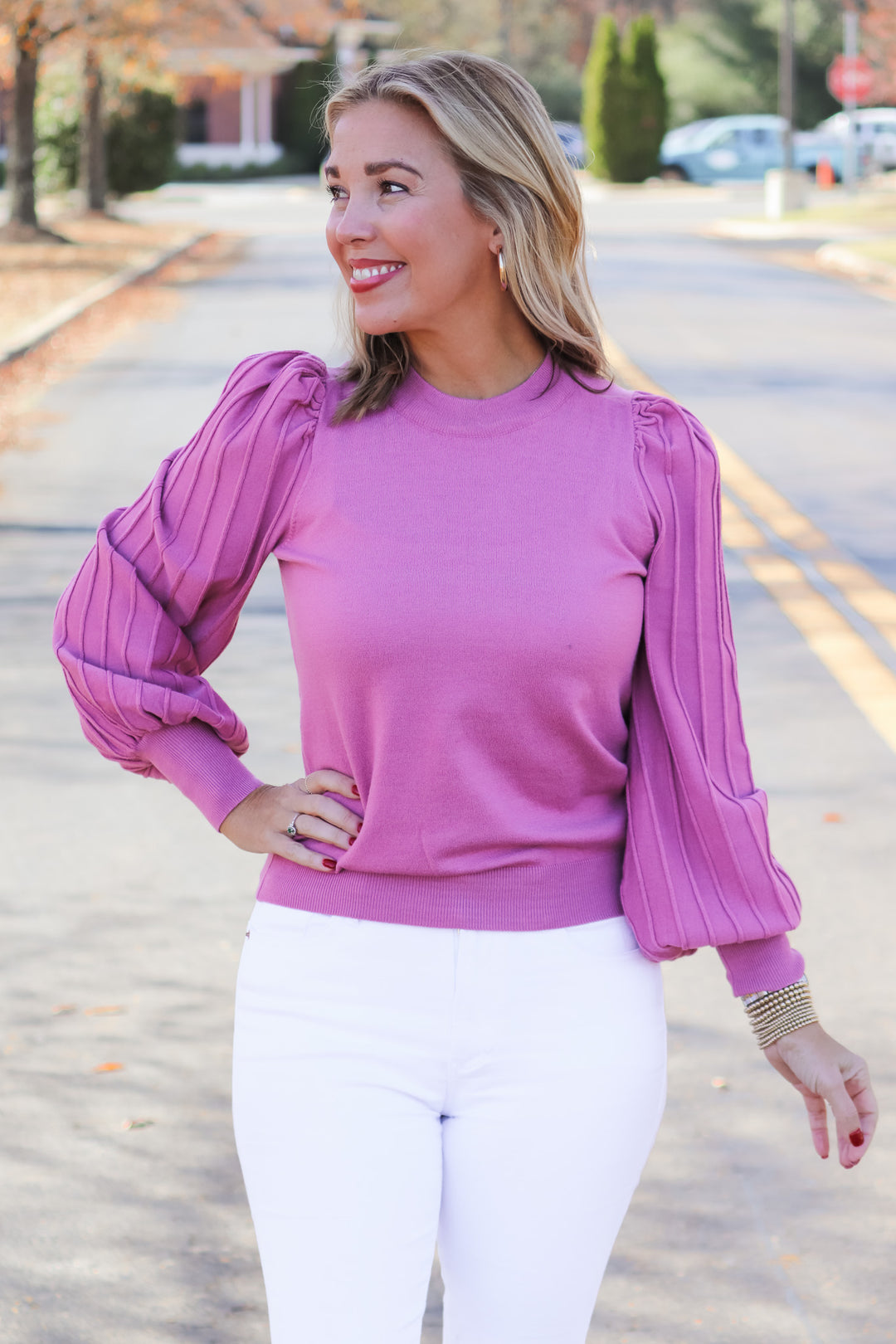 A blonde woman standing outside wearing a pink detailed long sleeve sweater and white jeans.