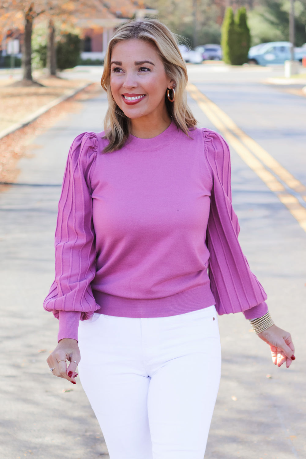 A blonde woman standing outside wearing a pink detailed long sleeve sweater and white jeans.