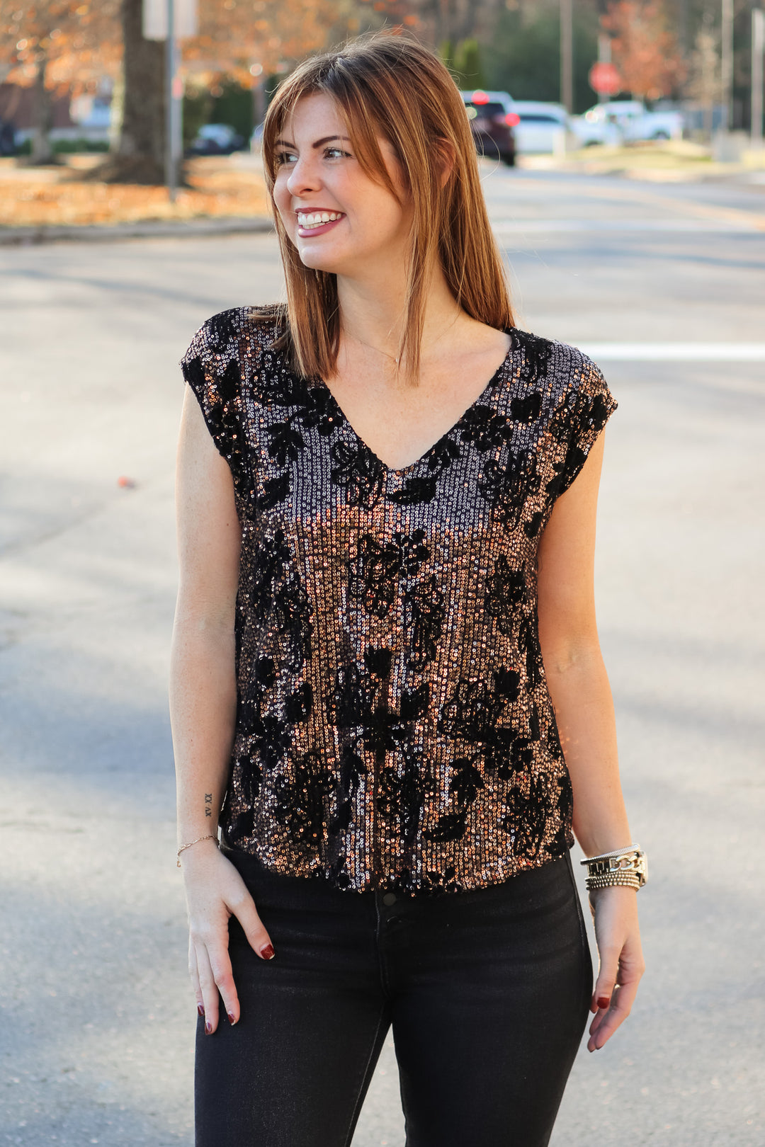 A brunette woman standing outside wearing a cap sleeve rose gold and black sequin top with a v neck and black jeans.