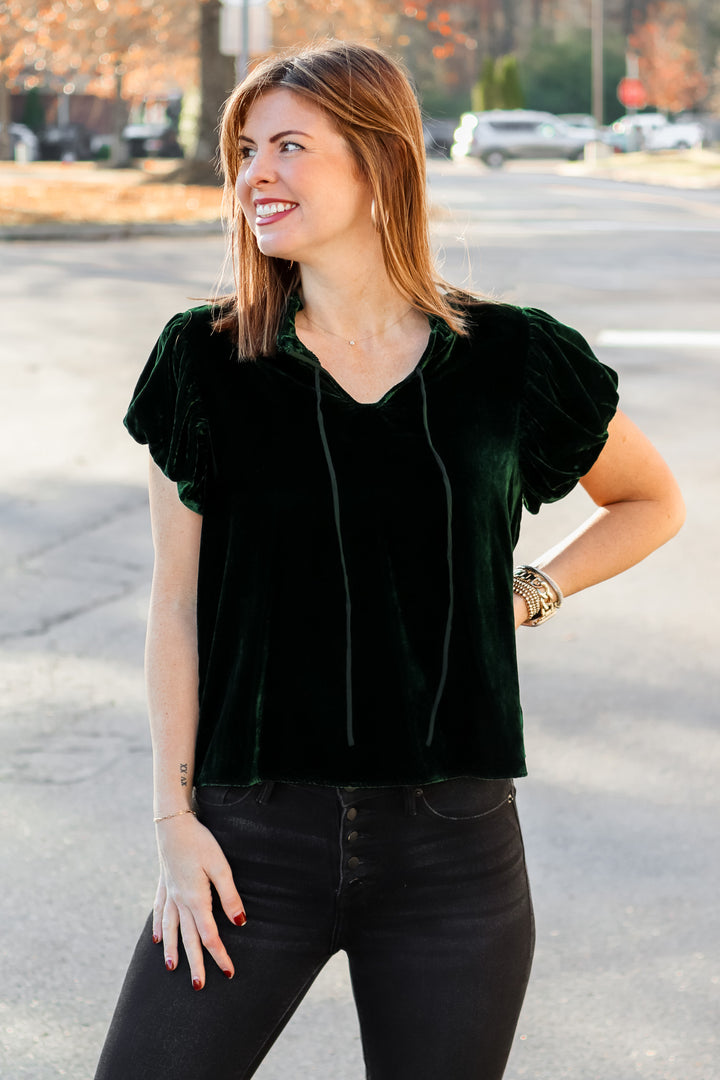 A brunette woman standing outside wearing a dark green velvet top with tie collar, short sleeves and black jeans.