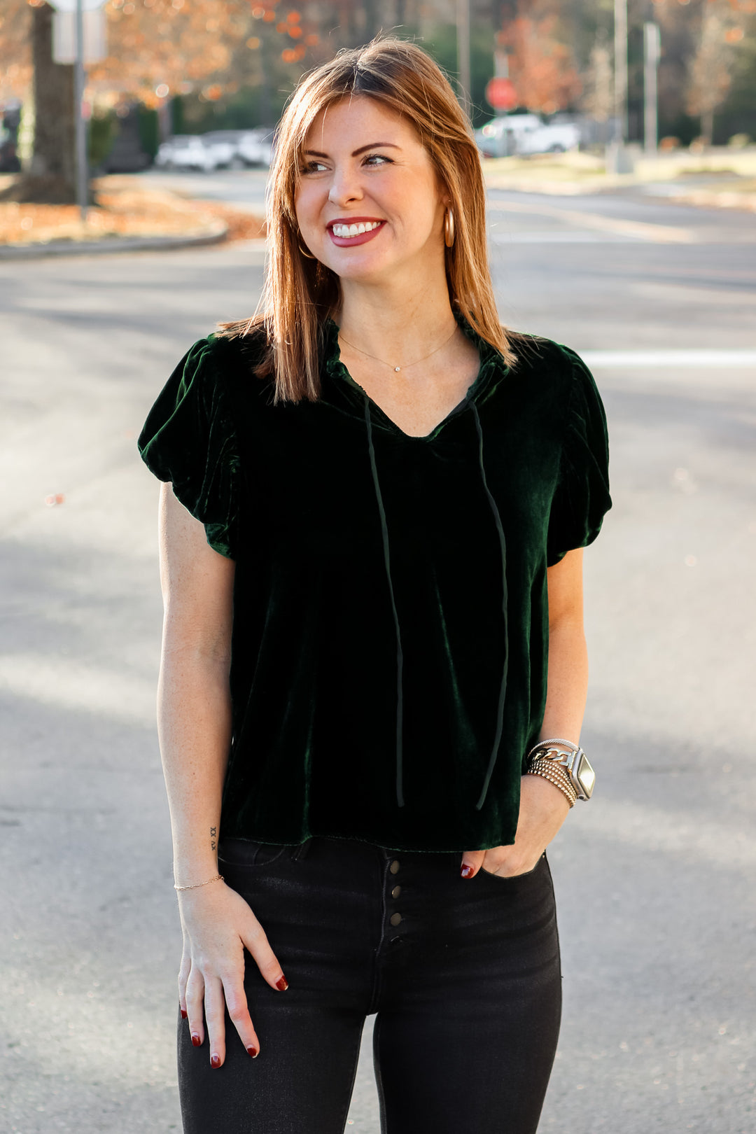 A brunette woman standing outside wearing a dark green velvet top with tie collar, short sleeves and black jeans.