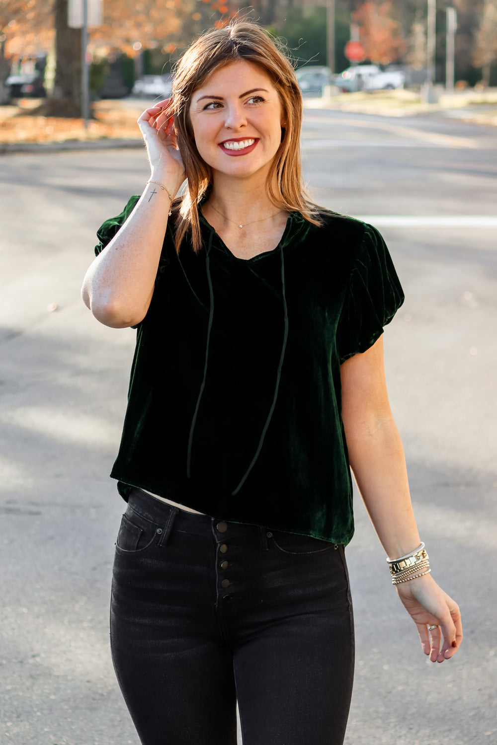 A brunette woman standing outside wearing a dark green velvet top with tie collar, short sleeves and black jeans.