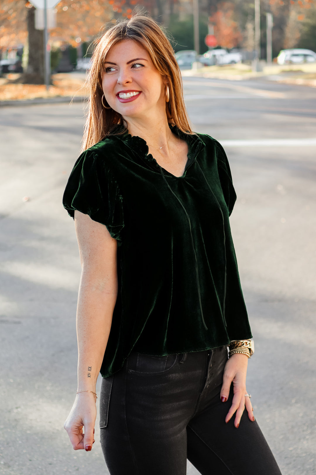 A brunette woman standing outside wearing a dark green velvet top with tie collar, short sleeves and black jeans.