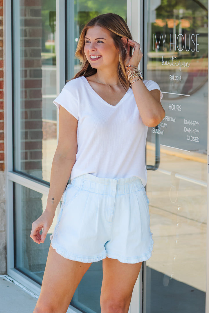 A brunette woman wearing a white shirt with cap sleeves and a v neck. She is standing in front of a shop.